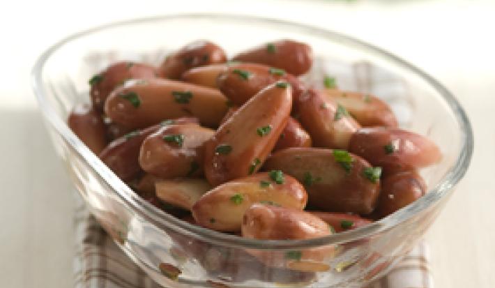 Fotografia em tons de marrom, com pote de vidro ao centro com pinhão sauteado salpicado de salsa, sobre guardanapo quadriculado nas cores branca e marrom, ao fundo ramo de salsa, tudo sobre bancada bege.