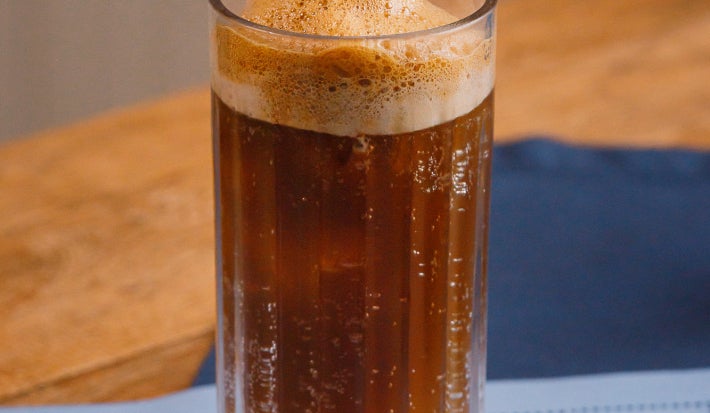 Copo alto com bebida escura gaseificada e espuma no topo, apoiado sobre tecido azul claro.