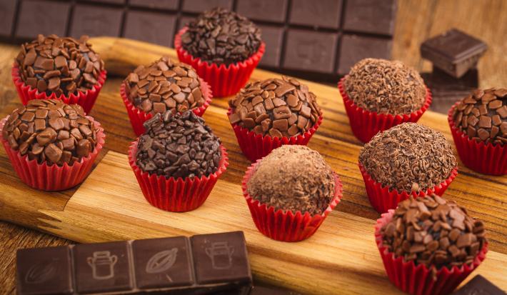 Fotografia em tons de vermelho em uma bancada de madeira com vários brigadeiros colocados em forminhas vermelhas em cima de uma tábua de madeira clara. Ao fundo, uma barra de chocolate meio amargo e com alguns pedaços de chocolate espalhados.