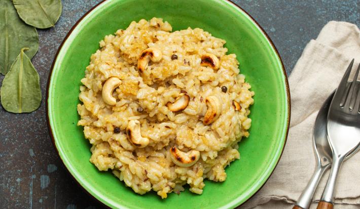 Fotografia vista de cima de um arroz crocante com castanha de caju dentro de um recipiente fundo verde, o qual está sobre uma mesa escura preta. Ao lado do recipiente tem um paninho e talheres por cima.