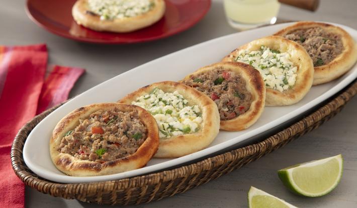 Foto da receita de Esfiha Aberta. Observa-se um recipiente branco de porcelana com 5 esfihas dispostas, intercaladas entre carne e queijo.