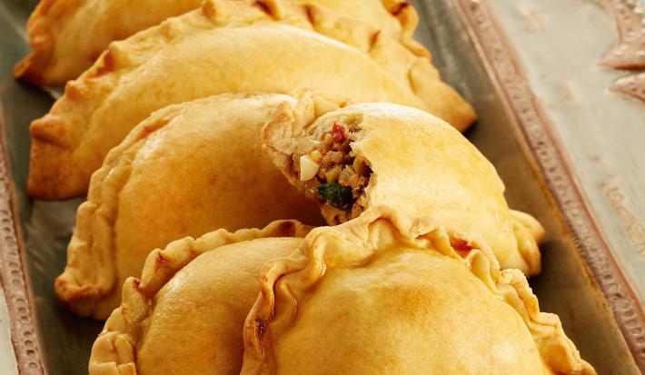 Fotografia em tons de amarelo e azul de uma bancada vista de cima. um recipiente cinza retangular Empanadas e abaixo do recipiente um pano nas cores azul, branco e rosa.contém varias