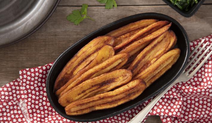 Fotografia em tons de vermelho em uma bancada de madeira, uma toalha vermelha com flores, um recipiente oval preto com várias bananas fritas dentro dele.
