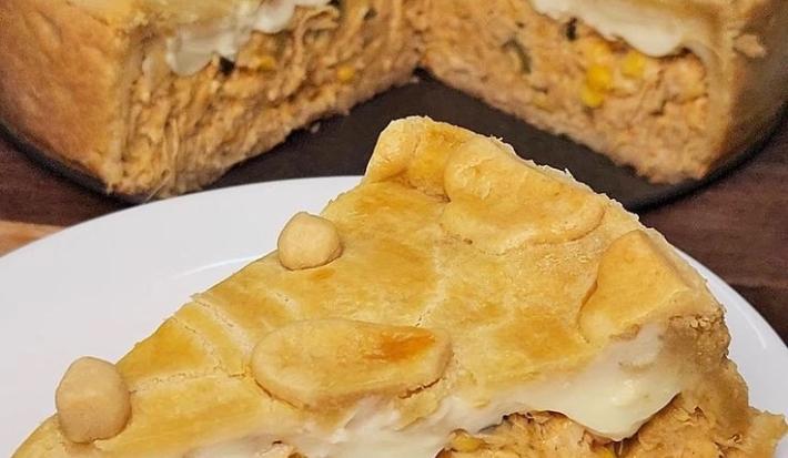 Fotografia de uma fatia de empadão de frango sobre um prato raso branco de vidro. No fundo tem o resto da torta quase inteira sobre o fundo da forma removível.