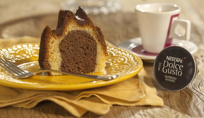 Fotografia em tons de marrom e amarelo de uma bancada de madeira com um paninho amarelo, sobre ele um prato redondo amarelo com uma fatia do bolo e um garfo. Ao fundo uma xícara branca com detalhes vermelhos e uma capsula de Dolce Gusto.