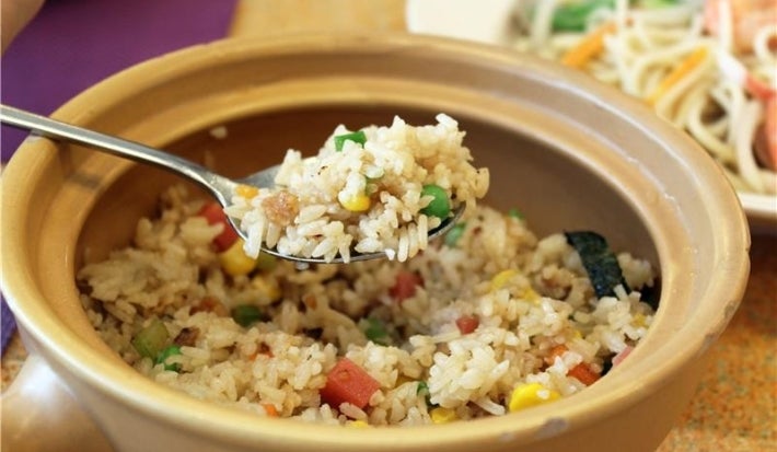 Em cima de uma mesa, tigela redonda com uma colher. Na colher, porção de arroz soltinho com ervilhas, cenouras picadas, uvas-passas brancas, pedaços de bacon.