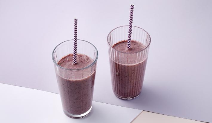 Fotografia em tons de roxo, em uma bancada de madeira, uma toalha lilás, um pano branco, dois copos de vidro altos com a vitamina de frutas vermelhas dentro deles, e um canudinho roxo dentro de cada copo.