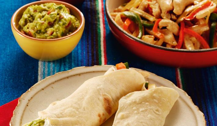 Fotografia em tons de vermelho, bege e azul, abaixo prato com duas fajitas sobre guardanapo vermelho, acima à direita panela vermelha com frango e legumes, à esquerda potinho amarelo com guacamole e outro com creme, tudo sobre toalha em tons de azul.