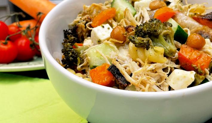 Fotografia em tons de verde com um recipiente fundo ao centro. Dentro do recipiente existe um macarrão com legumes e frango desfiado. Ao fundo existe um ramo de tomate cereja