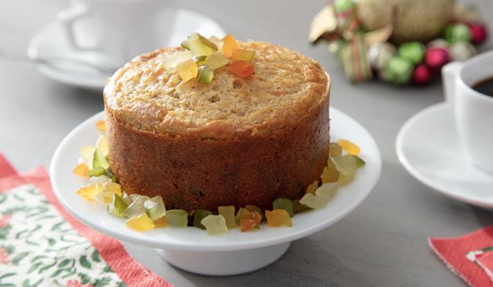 Fotografia em tons de amarelo em uma bancada de madeira de cor cinza. Ao centro, uma boleira contendo o panettone com frutas cristalizadas e uma toalha florida ao lado. Ao fundo, um pires com uma xícara branca de café e alguns enfeites natalinos.