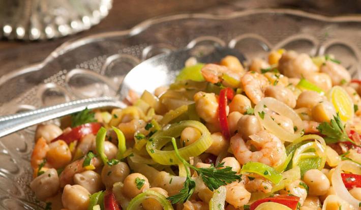 Fotografia em tons de bege e marrom, com salada de grã0-de-bico e legumes de cores verde e vermelha, dentro de saladeira de vidro com uma colher de servir, tudo sobre bancada de madeira.