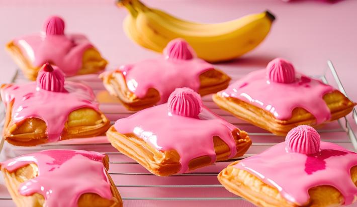 Fotografia de vários pasteis pequenos quadrados, recheados de banana e leite ninho, com cobertura de creme de nesquik. Os pasteis estão sobre uma grande branca, sobre uma mesa rosa.