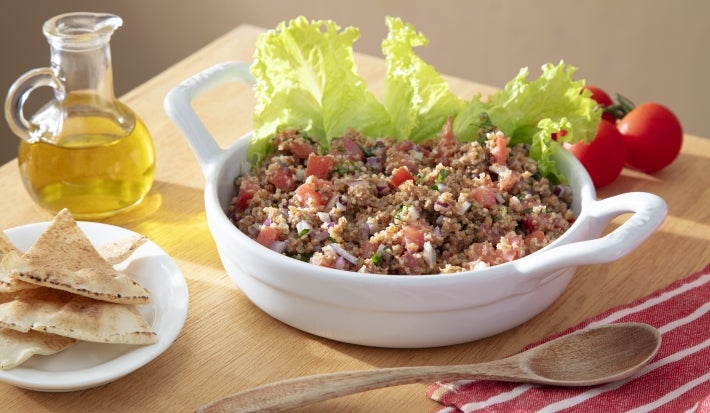 Fotografia em tons claros de um recipiente branco e fundo de vidro com uma tabule com folhas de alface. Ao redor do recipiente tem azeite, tomates pequenos, colher de madeira e pães sírios em um mini pratinho branco sobre uma mesa de madeira.