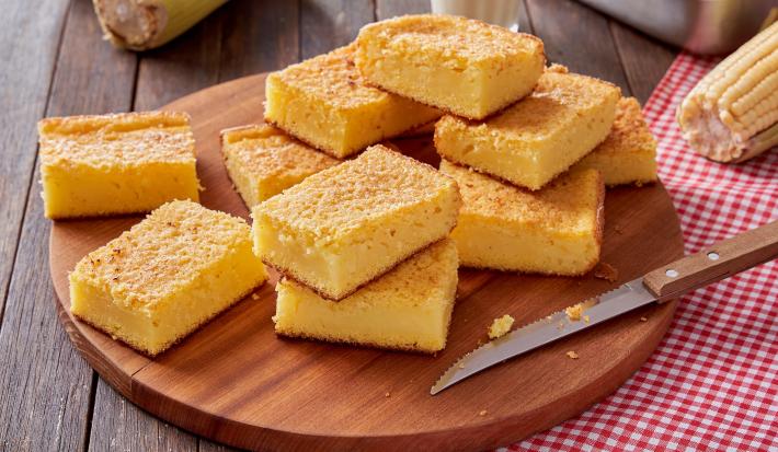 Foto da receita de bolo de milho cremoso, fatiado em pedaços sobre uma tábua de madeira sobre um pano quadriculado vermelho e branco. Na mesa há uma faca e espigas de milho