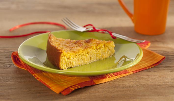 Em uma mesa contém um pano em tons laranja e vermelho, por cima, um prato na cor verde que comporta uma fatia de bolo. Ao lado do prato um garfo de metal e uma xicara laranja.