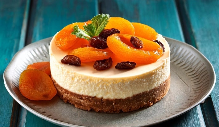 Fotografia de uma mini torta inteira sobre um prato cinza pequeno sore uma mesa de madeira azul. Sobre a torta tem damascos e frutas secas.