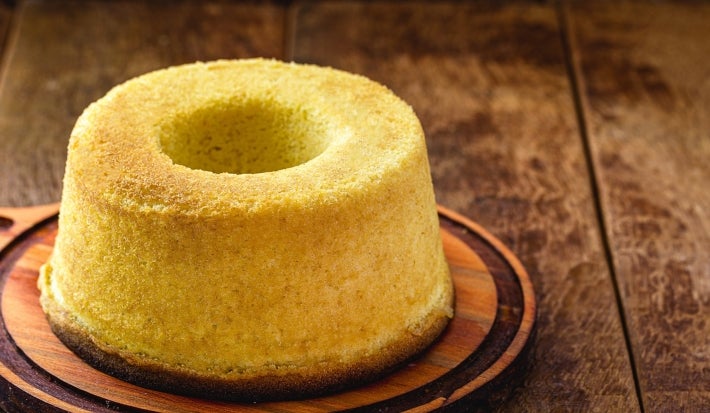 Fotografia de um bolo de milho recém-assado, com formato de bundt, dourado e macio, colocado em uma tábua de madeira. O fundo rústico.