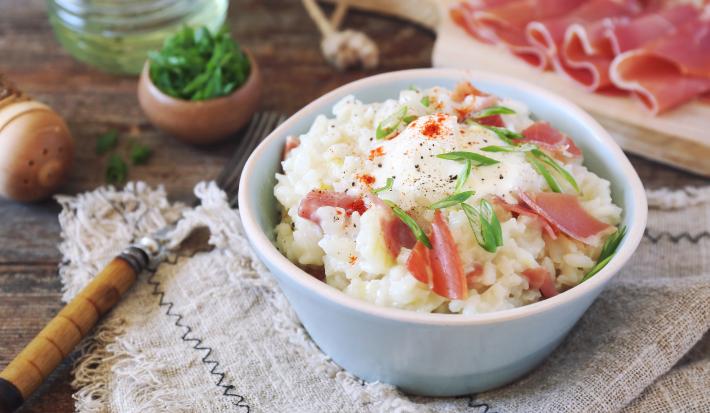 Fotografia em tons claros de um refratário branco pequeno e fundo sobre uma toalha de mesa, com arroz cremoso, peito de peru, lascas de presunto de Parma e cebolinha por cima.  Ao fundo, fatias do presunto sobre uma tábua de madeira e algumas decorações.