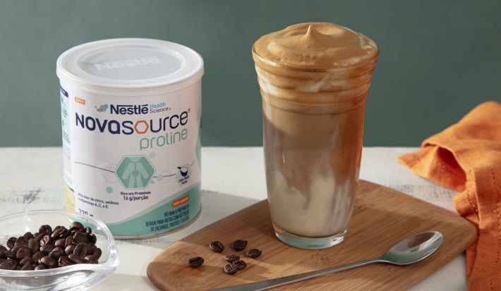 Foto da receita de café gelado cremoso servido em um copo de vidro alto sobre uma tábua de madeira com uma embalagem de novasource proline ao lado