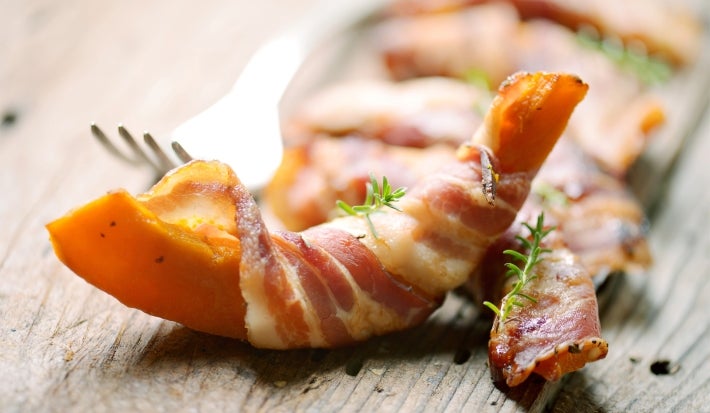 Em cima de uma mesa, há pedaços de abóboras cortadas em meia-lua enroladas com bacon.