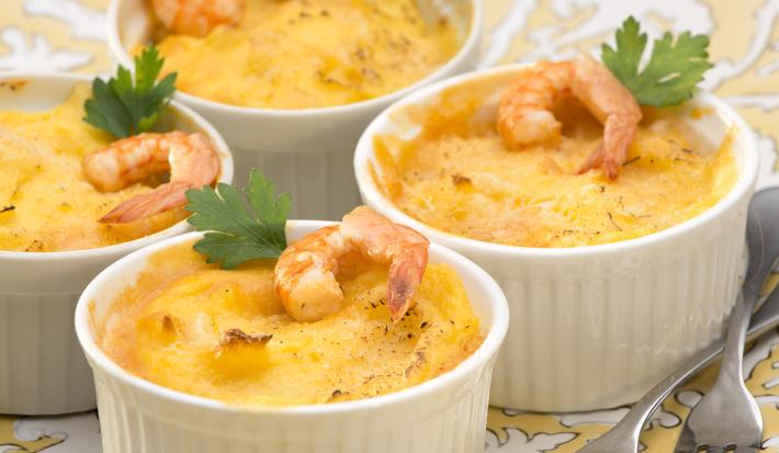 Fotografia em tons de branco e amarelo de uma bancada de madeira marrom, um paninho cinza e um prato amarelo com branco, sobre ele quatro recipientes redondo com o escondidinho de camarão decorados com um camarão e uma folha de salsinha.