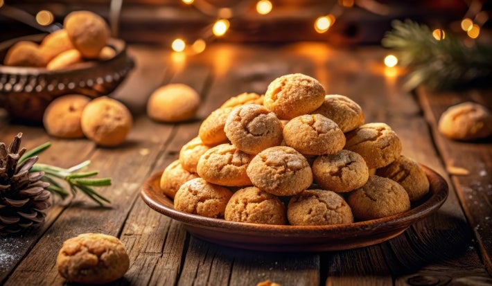 Fotografia em tons marrrom com um prato marrom ao centro. Em cima do prato existe vários biscoitos amaretti polvilhados com açúcar de confeiteiro.