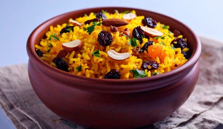 Tigela de porcelana redonda com porção de arroz amarelo e soltinho com amêndoas e uvas-passas pretas, decorado com folhas de cheiro-verde.
