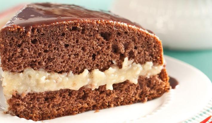Fotografia em tons de branco, marrom e azul. Um prato branco contém uma fatia de bolo de chocolate com recheio de coco.