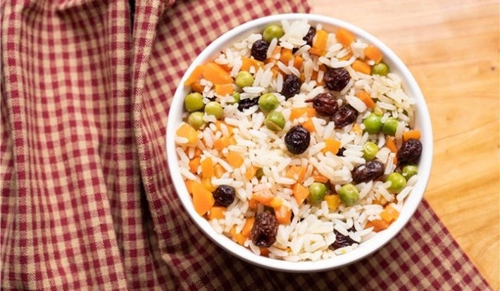 Em cima de uma mesa de madeira, há um pote branco  com uma porção colorida de arroz branco com uvas-passas, cenouras e ervilhas.
