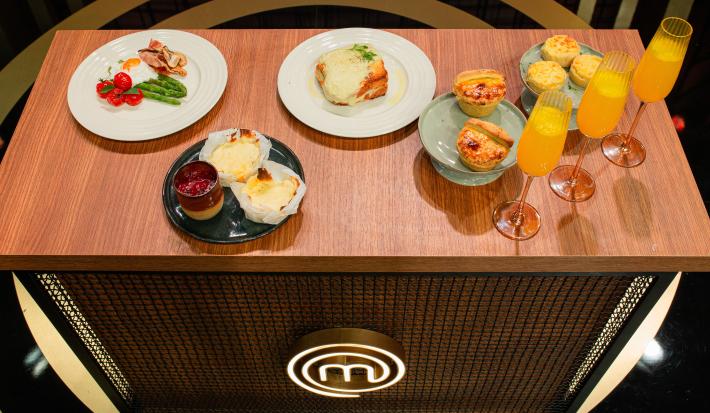 Fotografia de uma bancada de madeira com um brunch com quiche de alho poró, molho bechamel, legumes, ovo e empada, todos em pratos de cores diferentes, e três taças com suco.