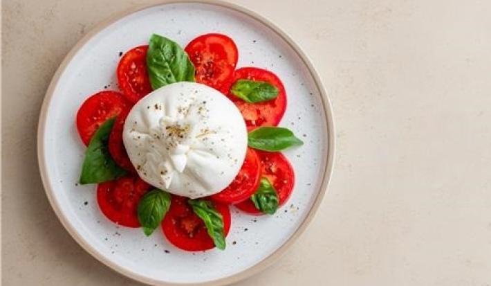 Fotografia vista de cima em tons claros de uma mesa de tom bege. Sobre a mesa tem um prato de cerâmica branco com uma burrata ao centro e rodelas de tomate em volta. Entre os tomates tem folhas de manjericão, sobre a burrata tem molho de manjericão.