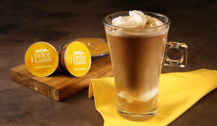 Fotografia em tons de amarelo em uma bancada de madeira escura, um paninho amarelo, uma xícara de vidro alta com o Latte Macchiato Dolce Gusto dentro, com doce de leite e decorado com chantilly. Ao fundo, cápsulas do café e um potinho com doce de leite.