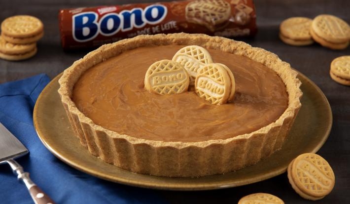 Foto de uma torta de doce de leite de cores marrom clara e escura. Ela está sobre um prato marrom, numa mesa com um tecido azul e uma espátula de bolo de aço inox. Há biscoitos Bono Doce de Leite espalhados e ao fundo um pacote de Biscoito Bono Foto de uma torta de doce de leite de cores marrom clara e escura. Ela está sobre um prato marrom, numa mesa com um tecido azul e uma espátula de bolo de aço inox. Há biscoitos Bono Doce de Leite espalhados e ao fundo um pacote de Biscoito Bono