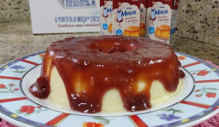 Imagem de um Pudim Romeu e Julieta sobre um prato colorido, numa bancada de cozinha com algumas embalagens do produto Moça Pra Toda a Família ao fundo