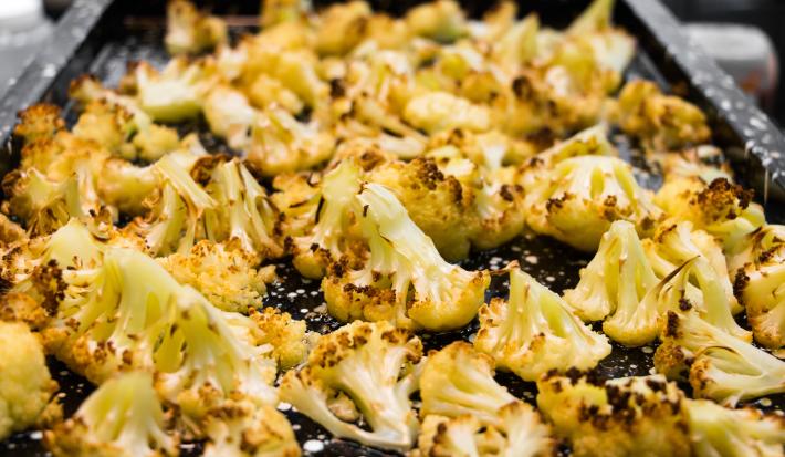 Fotografia em tons de preto com uma travessa grande ao centro. Em cima da travessa existe vários ramos de couve-flor gratinados e temperados com especiarias.