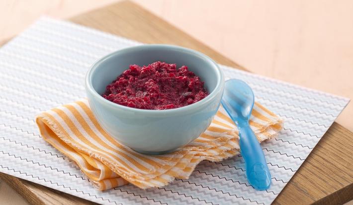 Fotografia em tons de roxo em uma bancada de madeira de cor marrom. Ao centro, uma tábua de madeira contendo um pano azul e um pano laranja, em cima há um recipiente contendo o purê. Ao lado, há uma colher azul.