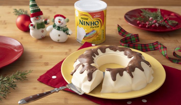 Fotografia em tons de vermelho e branco de uma bancada de madeira, um paninho vermelho e um prato amarelo com o bolo de sorvete com calda de chocolate em cima. Ao fundo decoração de natal, com mini bonecos de neve e uma lata de NINHO.