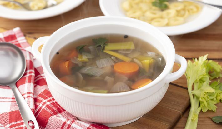 Fotografia em tons de vermelho em uma bancada de madeira, um pano xadrez vermelho, uma panela branca funda com alças laterais com o caldo de legumes dentro dela. Ao lado, uma concha e dois pratos brancos fundo com sopa de capelleti com o caldo de legumes.