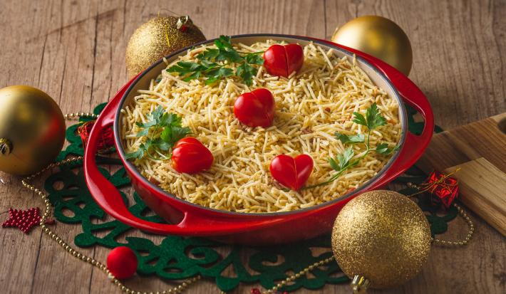 Fotografia em tons de vermelho em uma bancada de madeira, um pano redondo natalino verde, um prato vermelho redondo grande e fundo com a salada de salpicão coberta com batata palha e tomate-cereja dentro dele. Ao lado, enfeites de Natal.