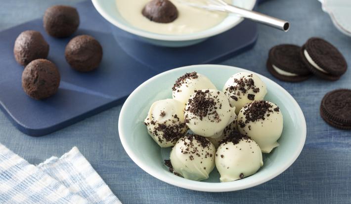 Foto da receita de Trufa de Negresco. Observa-se um recipiente verde menta com as tufas dentro cobertas de chocolate branco. Atrás, uma tábua azul com as trufas sem cobertura e a foto está decorada com biscoitos Negresco.