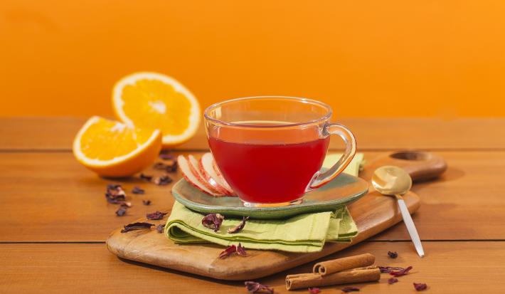 Fotografia em tons de laranja em uma bancada de madeira e um fundo laranja. Ao centro, uma tábua de madeira, um paninho verde e uma xícara de vidro com o chá de hibisco dentro dele. Ao fundo, laranja e canela em pau espalhados pela bancada.