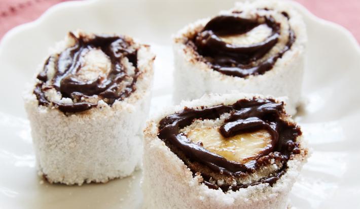 Fotografia em tons de vermelho em uma bancada de madeira clara, um pano vermelho, um prato branco redondo com três rolinhos de tapiocas com banana e chocolate.