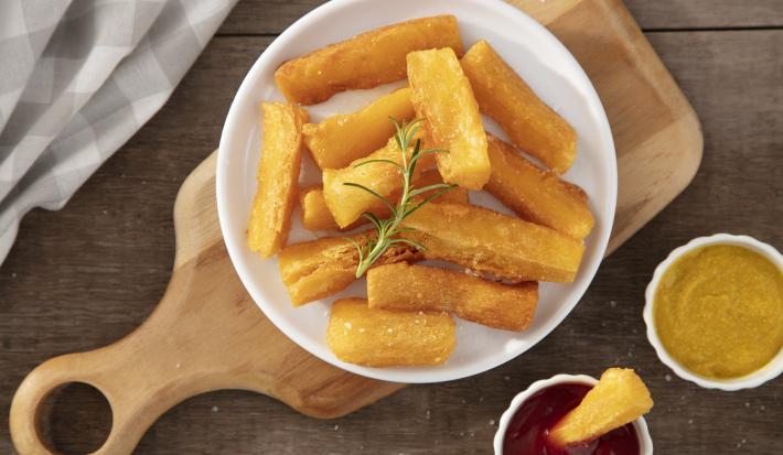 Imagem vista de cima de um prato branco com mandioca frita sobre uma tábua de madeira. Ao lado, um potinho de ketchup e um de mostarda.