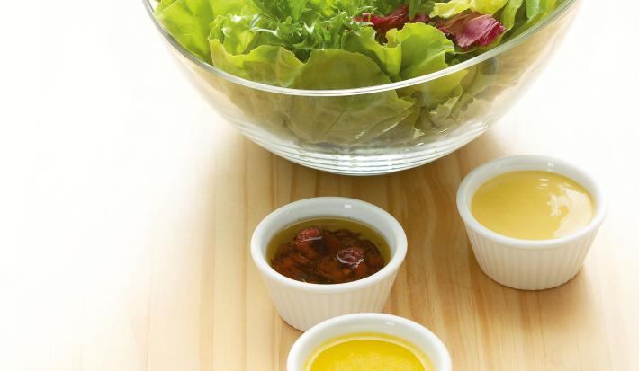 Fotografia em tons de verde em uma mesa de madeira clara com um bowl de vidro com salada de alface e rúcula e ao lado, quatro potinhos brancos com os molhos de morango, mostarda, hortelã e gengibre.