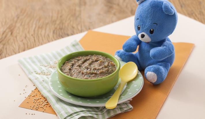 Fotografia em tons de laranja e verde de uma bancada de madeira com paninho verde listrado, um potinho verde em cima de um pratinho verde de bolinha com a sopinha de carne, couve e cenoura. Ao lado, uma colher pequena amarela e um ursinho de pelúcia azul.