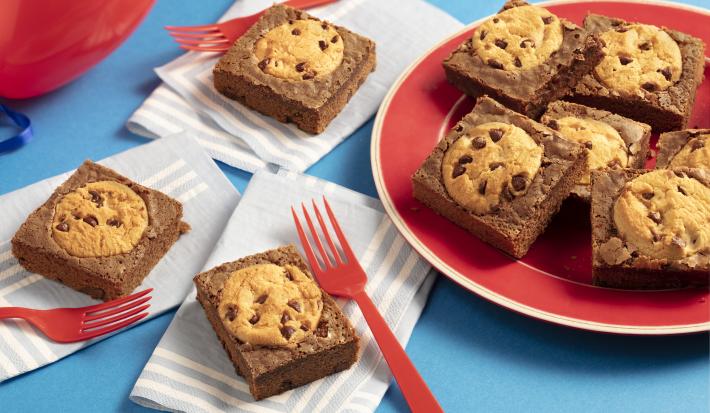 Fotografia em tons de azul e vermelho de uma bancada azul com paninhos azul claro com listras brancas e sobre eles pedaços de brownie com cookies e garfos vermelhos. Ao lado um prato vermelho e sobre eles pedaços de brownie com cookies.