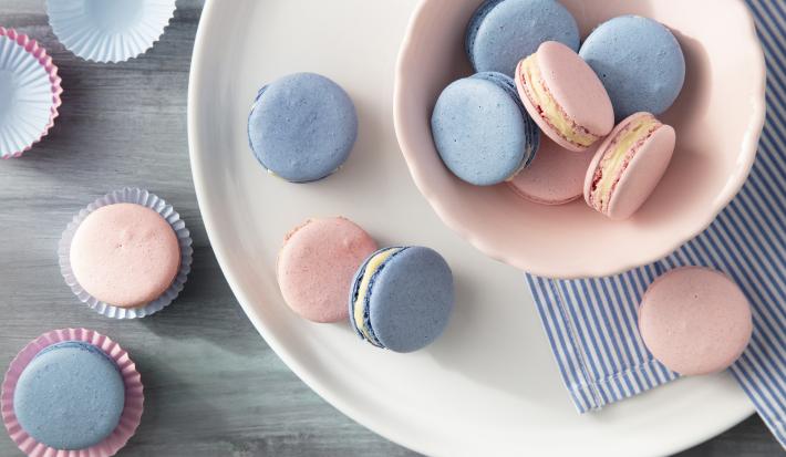 Imagem vista de cima de macarons rosa e azul em um pote rosa claro e ao lado, macarons em forminhas de papel.