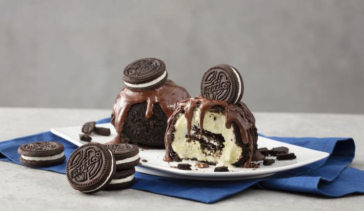fotografia em tons de cinza, azul branco e marrom de uma bancada cinza. Contém um pano azul com um prato retangular branco que contém duas bolas de sorvete, por cima calda de chocolate e biscoitos.