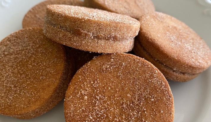 Fotografia em tons de marrom com um prato branco ao centro. Dentro do prato existe algumas unidades de biscoitos de cor caramelo recheados com doce de leite e cobertos com açúcar.