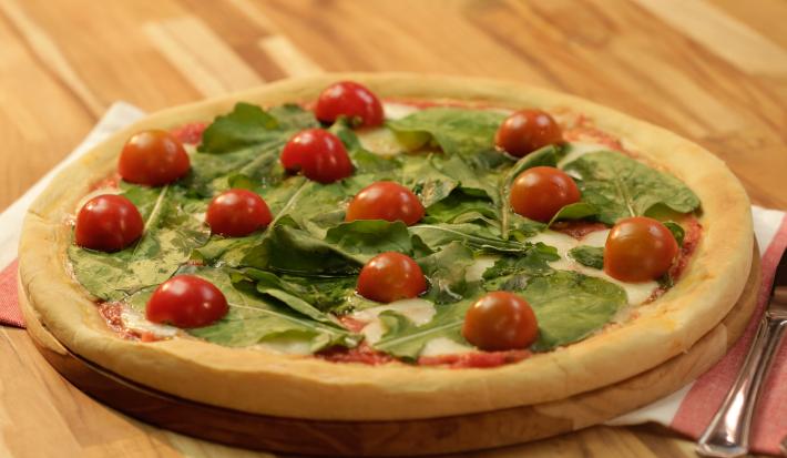 Fotografia em tons de dourado, verde e vermelho, ao centro pizza com folhas verdes e tomate-cereja sobre tábua redonda e guardanapo branco e vermelho, com talher ao lado, tudo sobre bancada de madeira em tons marrom.